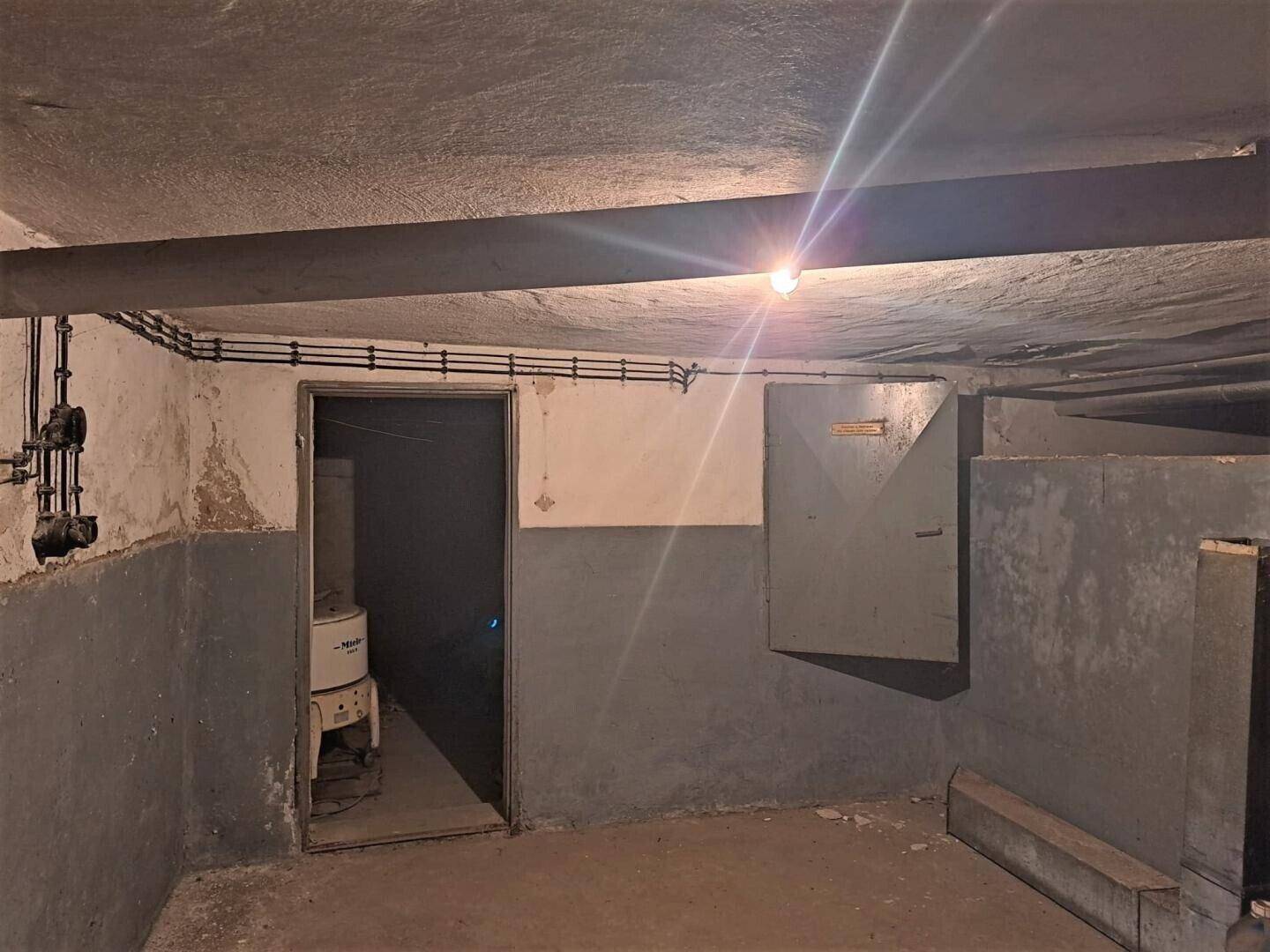 Dunkler Kellerraum mit Betonwänden, sichtbaren Rohren und einfacher Beleuchtung.