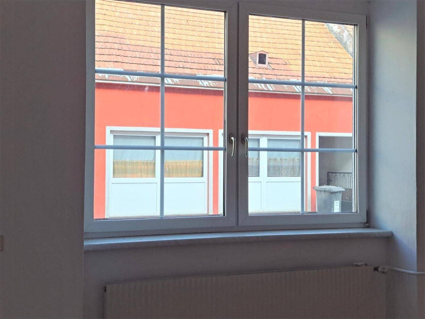 Fenster mit Blick auf ein gegenüberliegendes Gebäude mit rotem Dach und weiteren Fenstern.