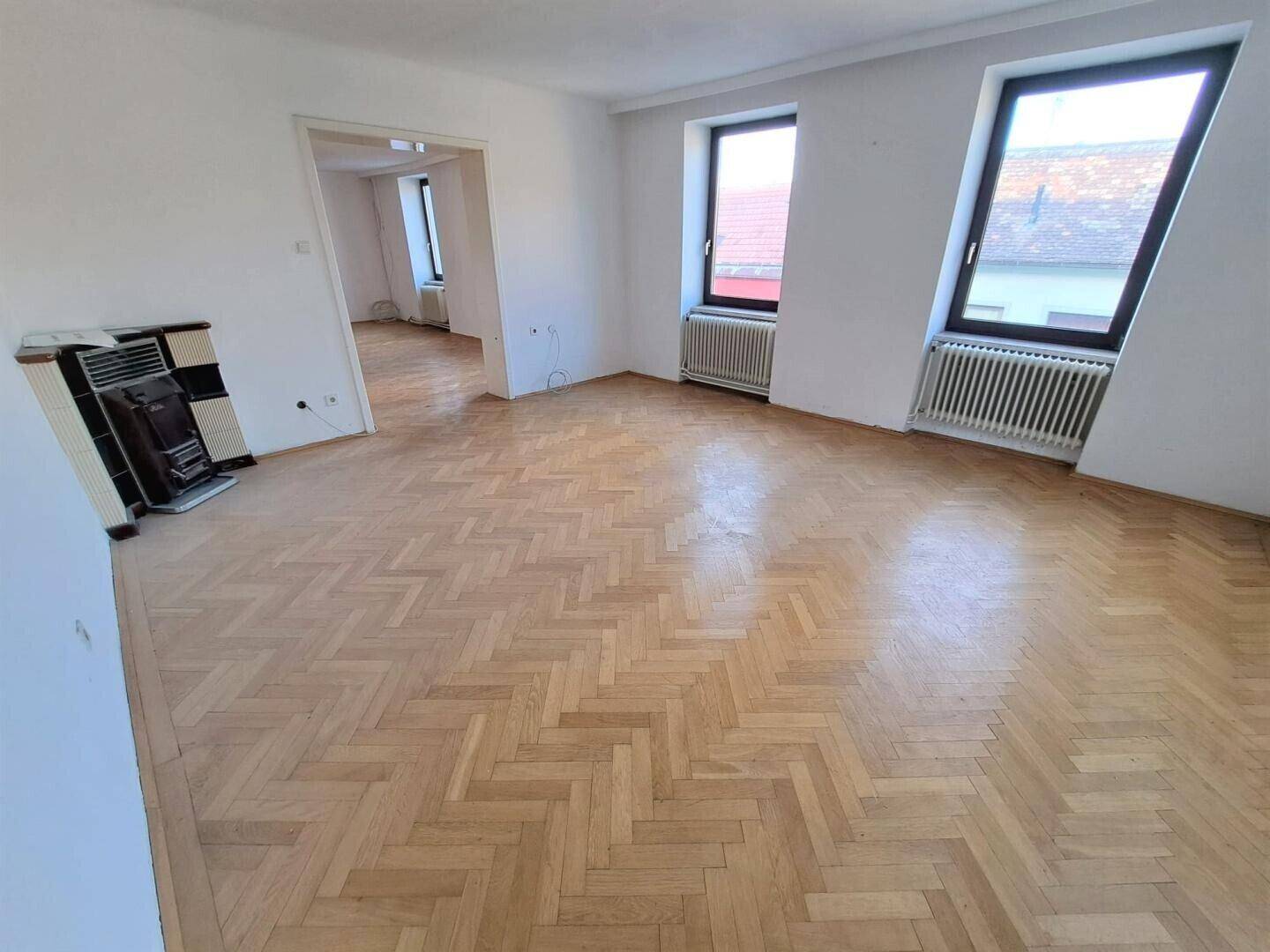 Geräumiges Wohnzimmer mit Fischgrätparkett und traditionellem Kachelofen, hell durch zwei Fenster.