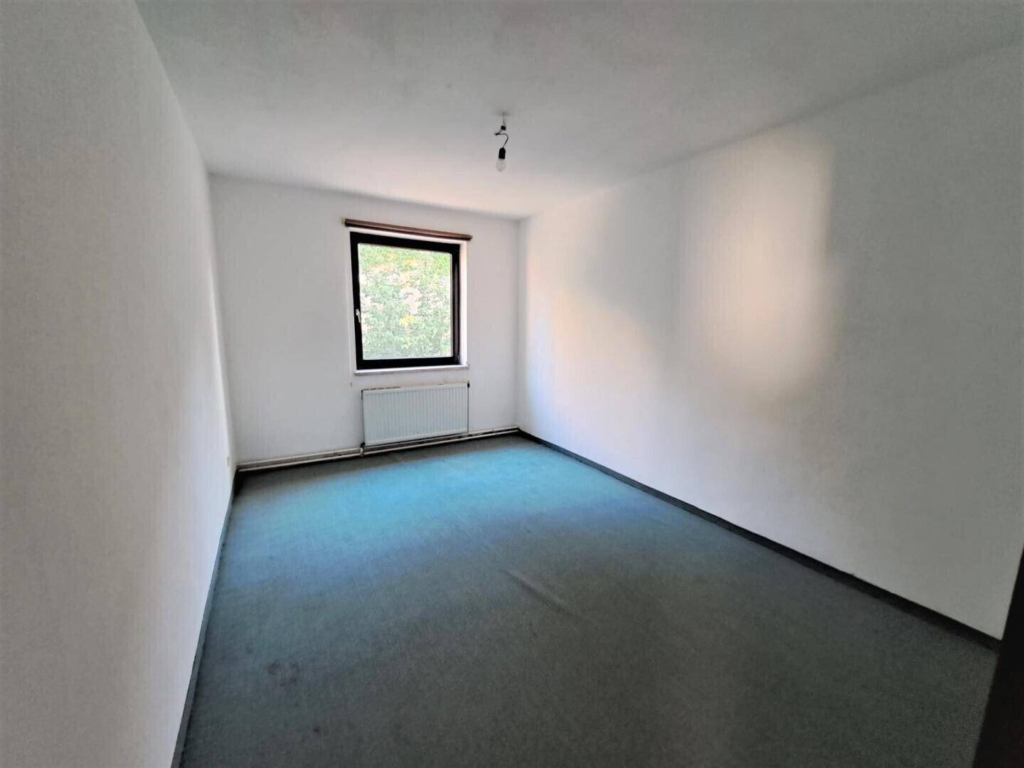 Schlichtes Schlafzimmer mit Teppichboden und einem Fenster, das Tageslicht hereinlässt.
