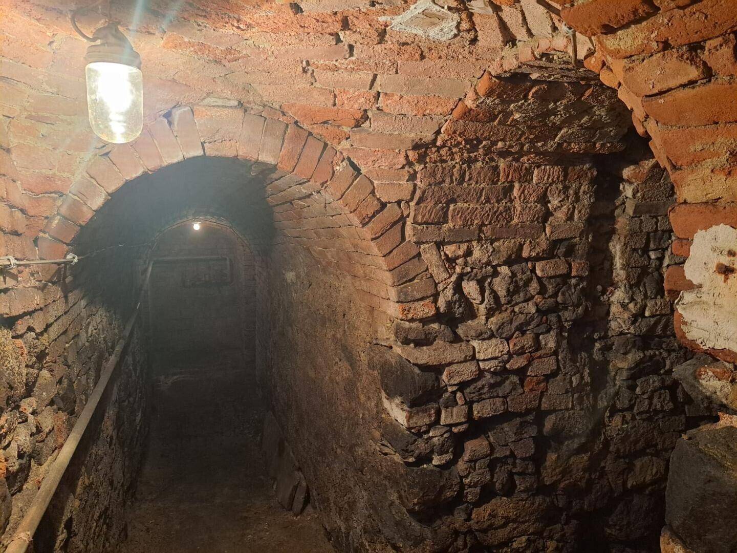 Historischer Keller mit Ziegelgewölben und freiliegenden Ziegelwänden, beleuchtet von einer einzelnen Glühbirne.
