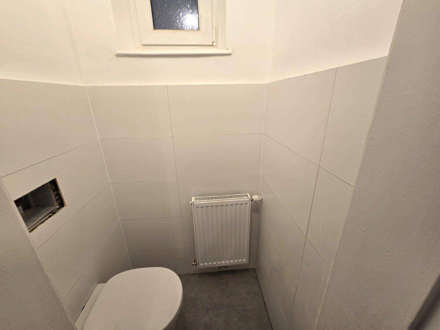Kleines Gäste-WC mit Wand-WC, Heizkörper und einem Fenster für natürliche Belüftung.