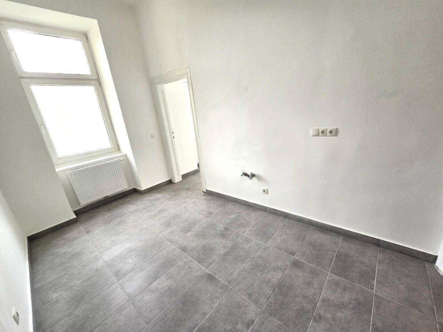 Leerer Raum mit grauen Fliesen, weiß gestrichenen Wänden und einem Fenster mit Heizkörper.