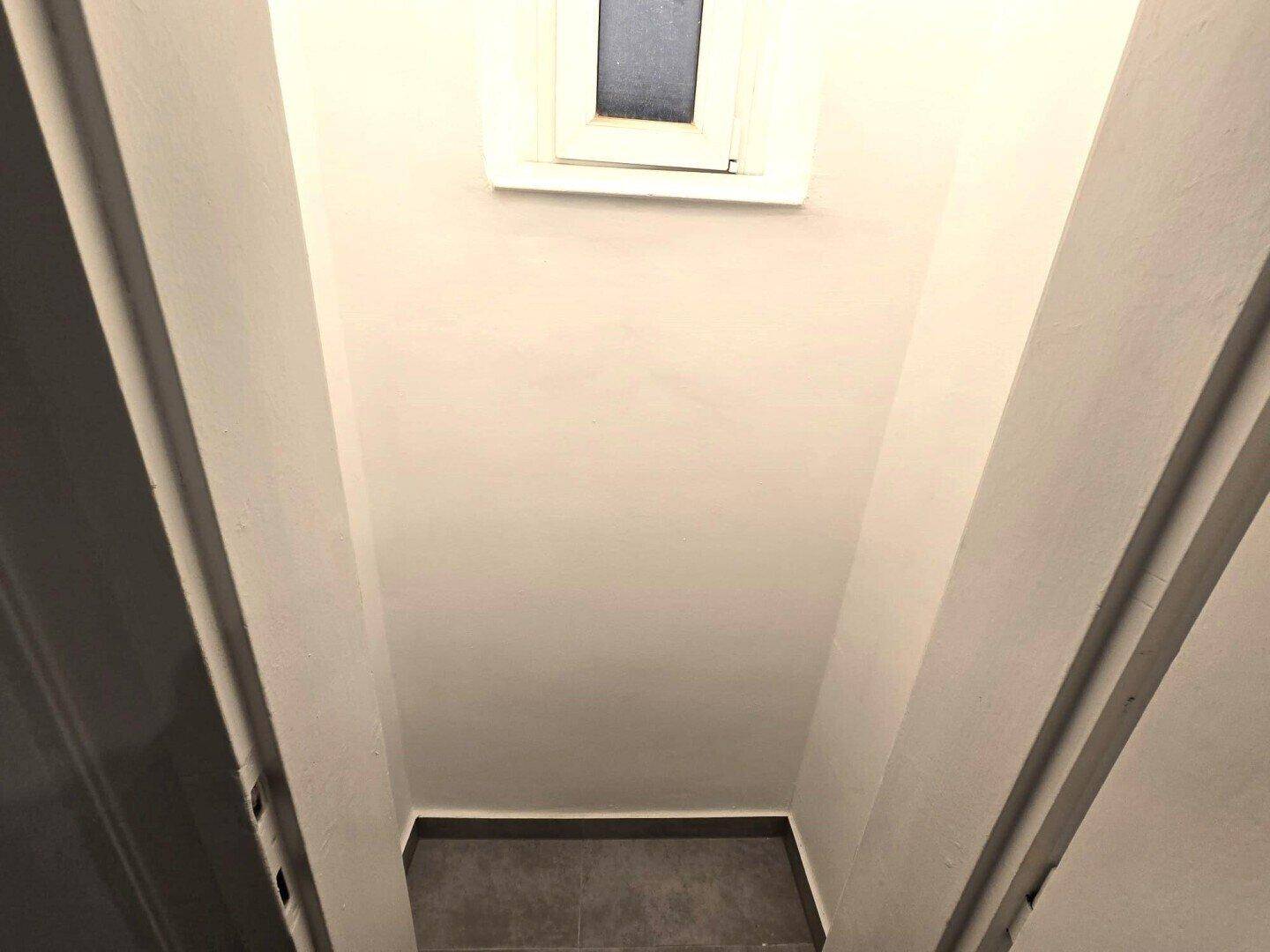 Schmaler Flur mit hellen Wänden und einem kleinen Fenster, das Tageslicht spendet.