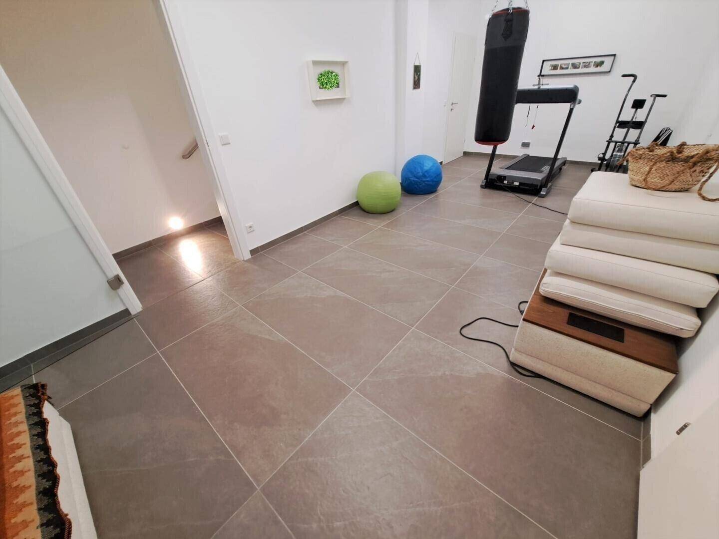 Heller Fitnessraum mit Fliesenboden und Zugang zu weiteren Räumen.