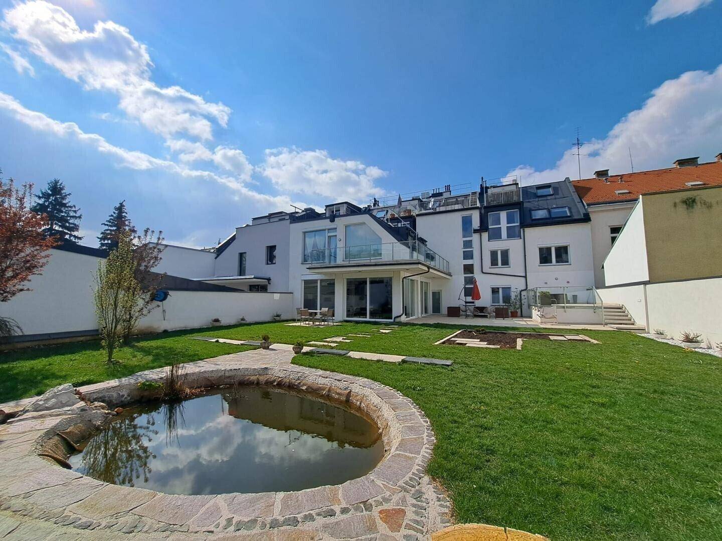 Weitläufiger Garten mit Teich, Rasenfläche und moderner Wohnanlage im Hintergrund.