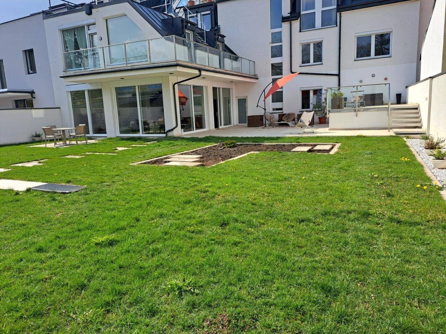 Gepflegter Garten mit Rasenfläche, kleiner Terrasse und modernem Gebäude im Hintergrund.