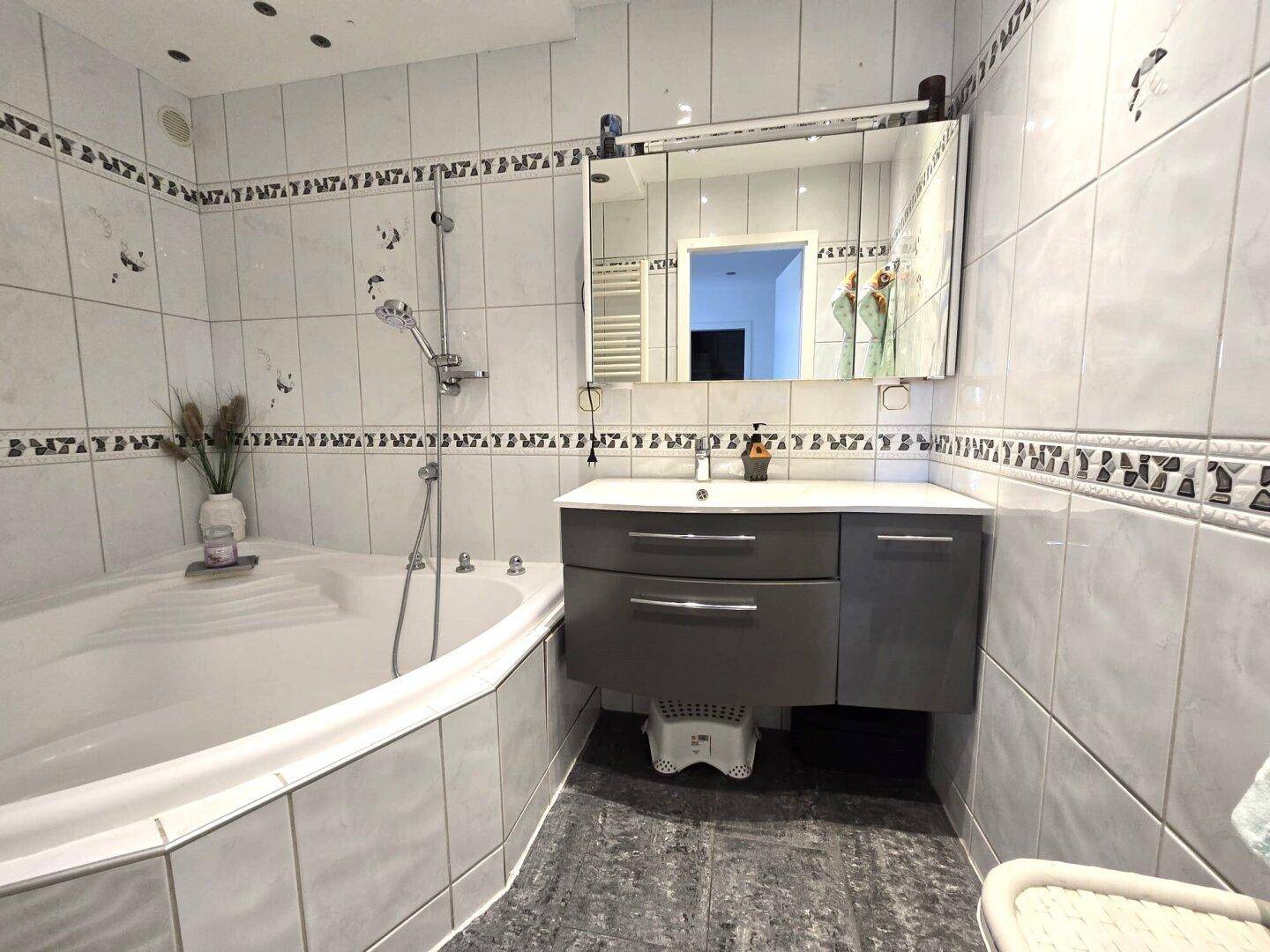 Badezimmer mit Eckbadewanne, Waschtisch und Spiegel, alles in hellen Fliesen gehalten.