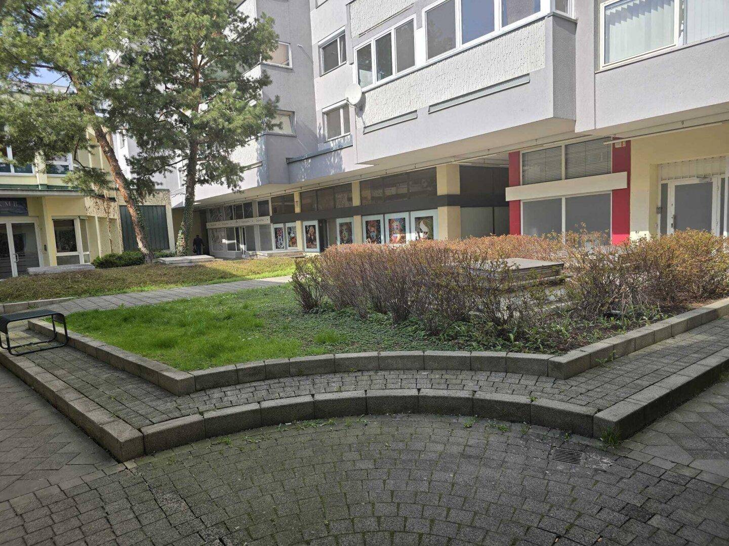 Grüner Innenhof mit gepflasterten Wegen und Sitzgelegenheiten, umgeben von Gebäuden.