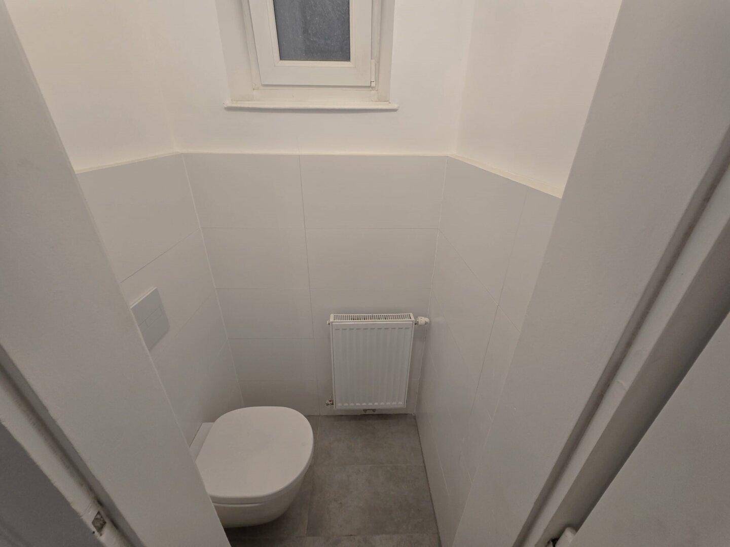 Kleines Gäste-WC mit Wand-WC, Heizkörper und einem Fenster für natürliche Belüftung.