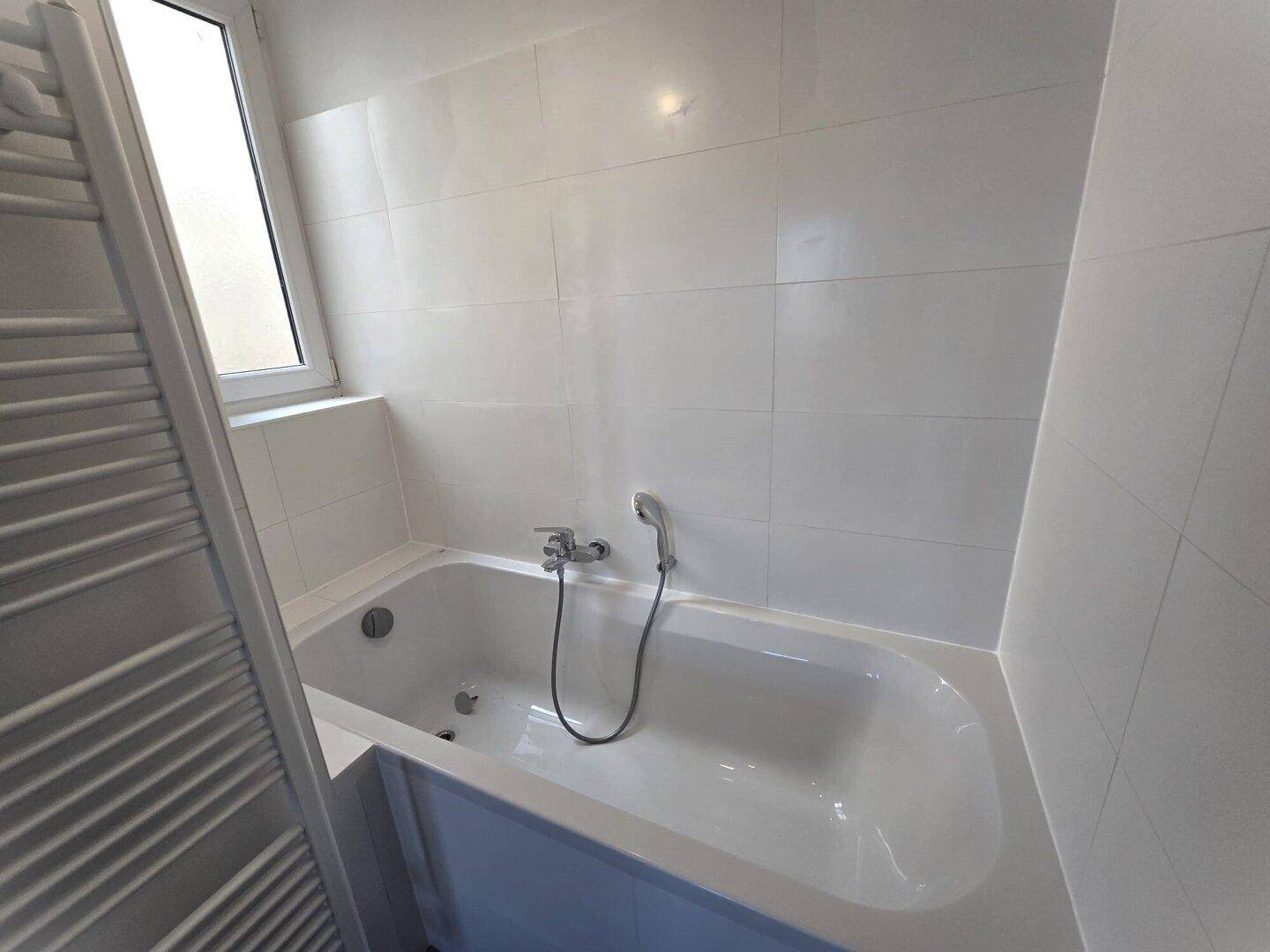Weiße Badewanne mit Duscharmatur und Fenster im hell gefliesten Badezimmer.