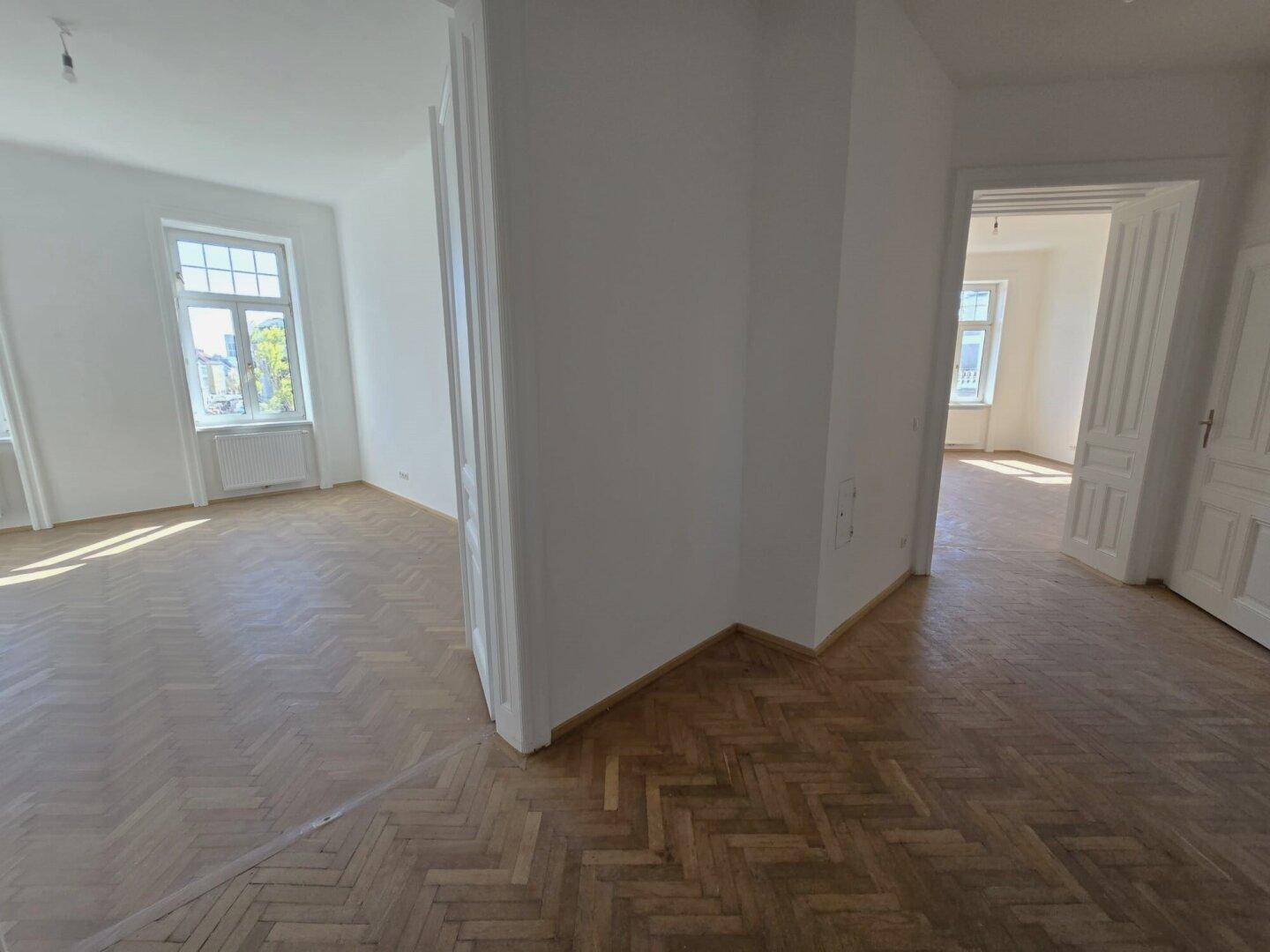 Zwei helle, leere Räume mit Fischgrätparkett und weißen Wänden, verbunden durch einen breiten Durchgang.