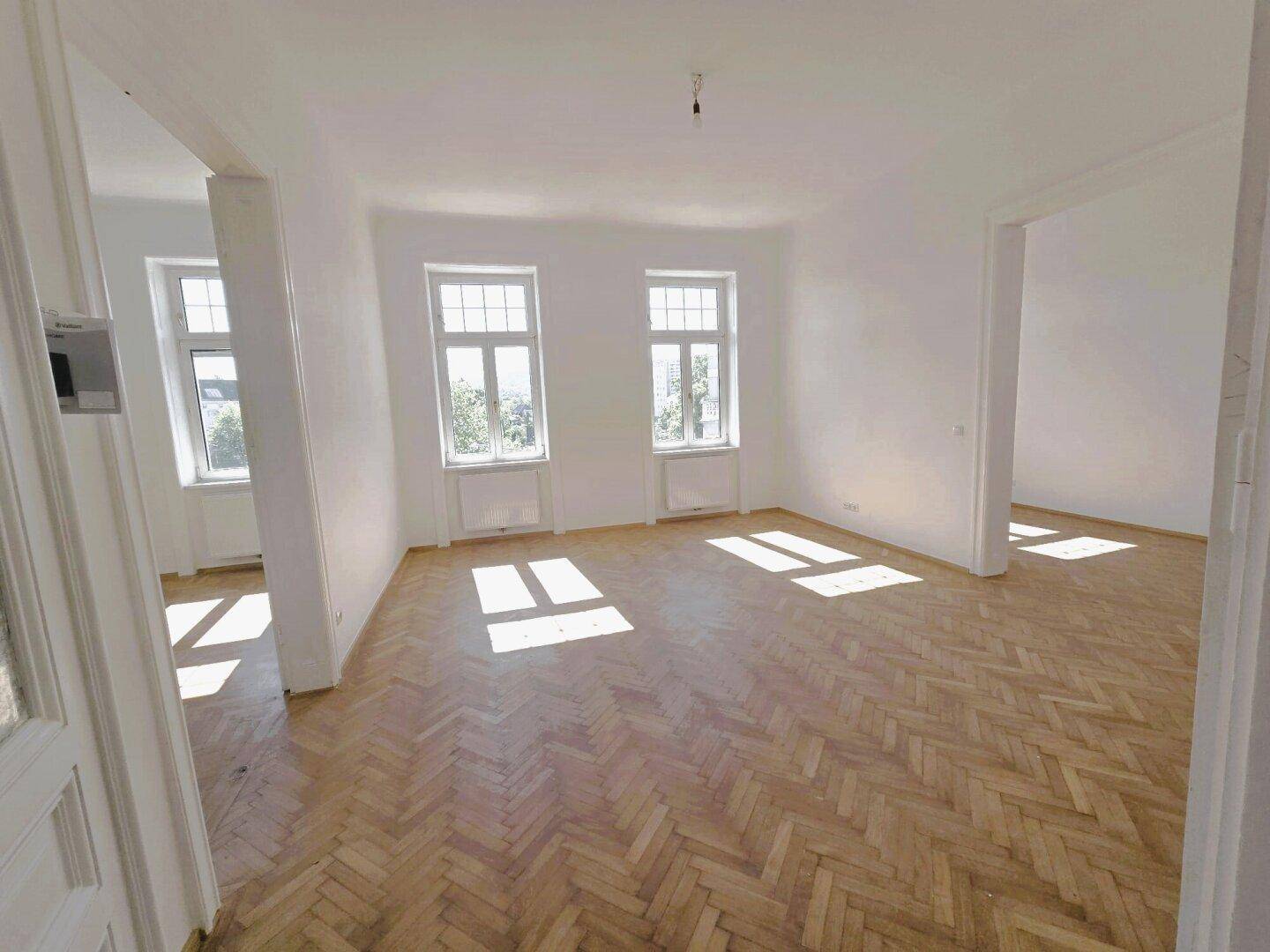 Helles, leeres Zimmer mit Fischgrätparkett und großen Fenstern, durch die Sonnenlicht einfällt.