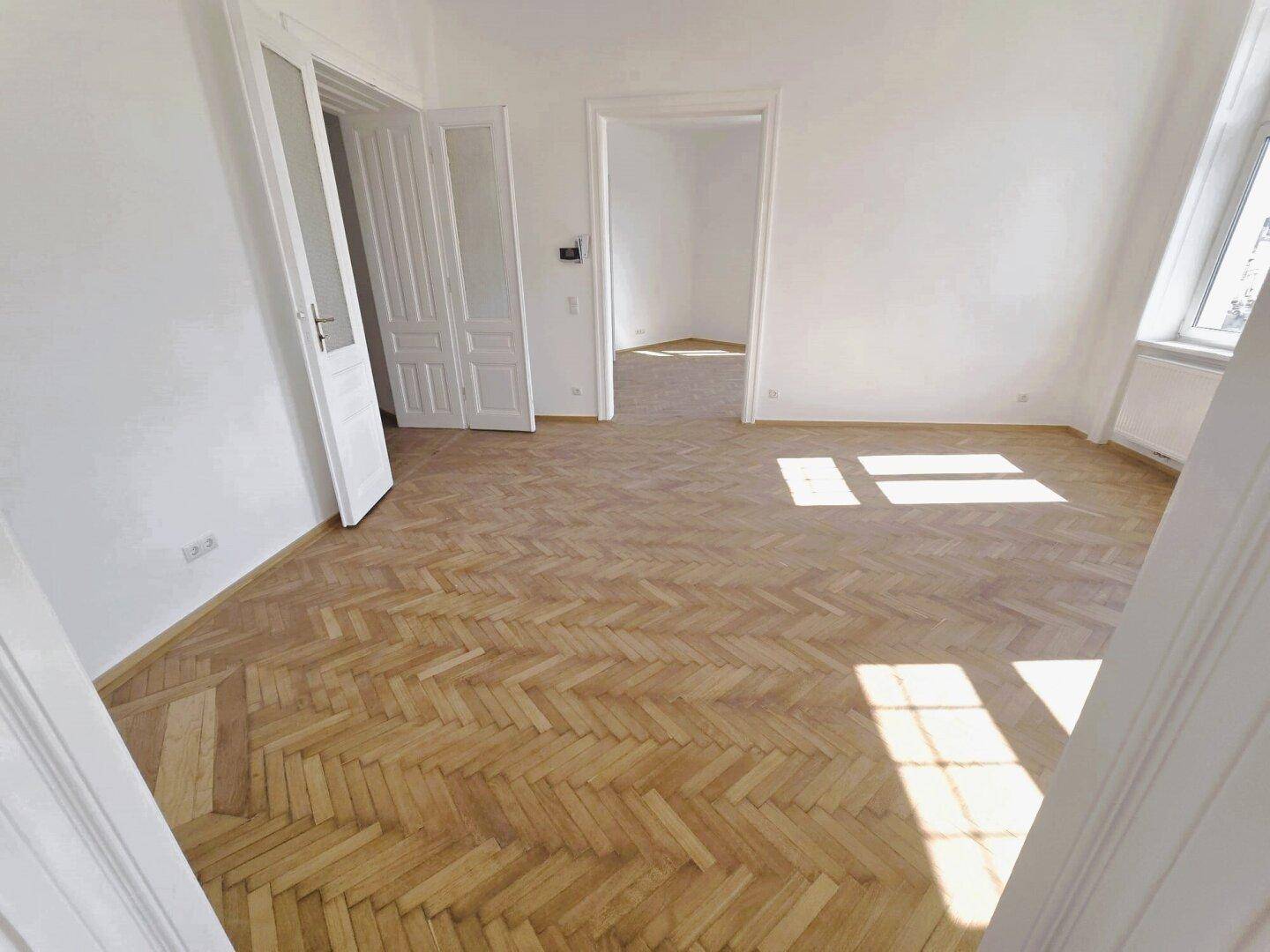 Geräumiges, helles Zimmer mit Fischgrätparkett und offenen Doppeltüren, die zu einem weiteren Raum führen.