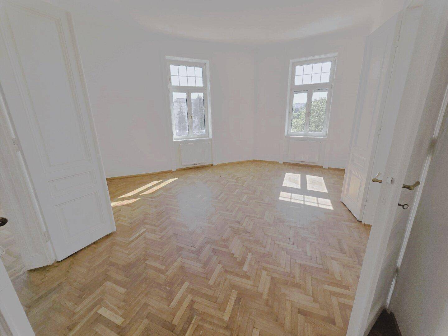 Helles, leeres Zimmer mit Fischgrätparkett und zwei großen Fenstern, die viel Tageslicht hereinlassen.
