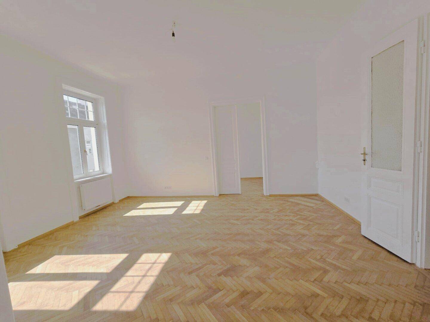 Helles, leeres Zimmer mit Fischgrätparkett, einem großen Fenster und einer offenen Tür.