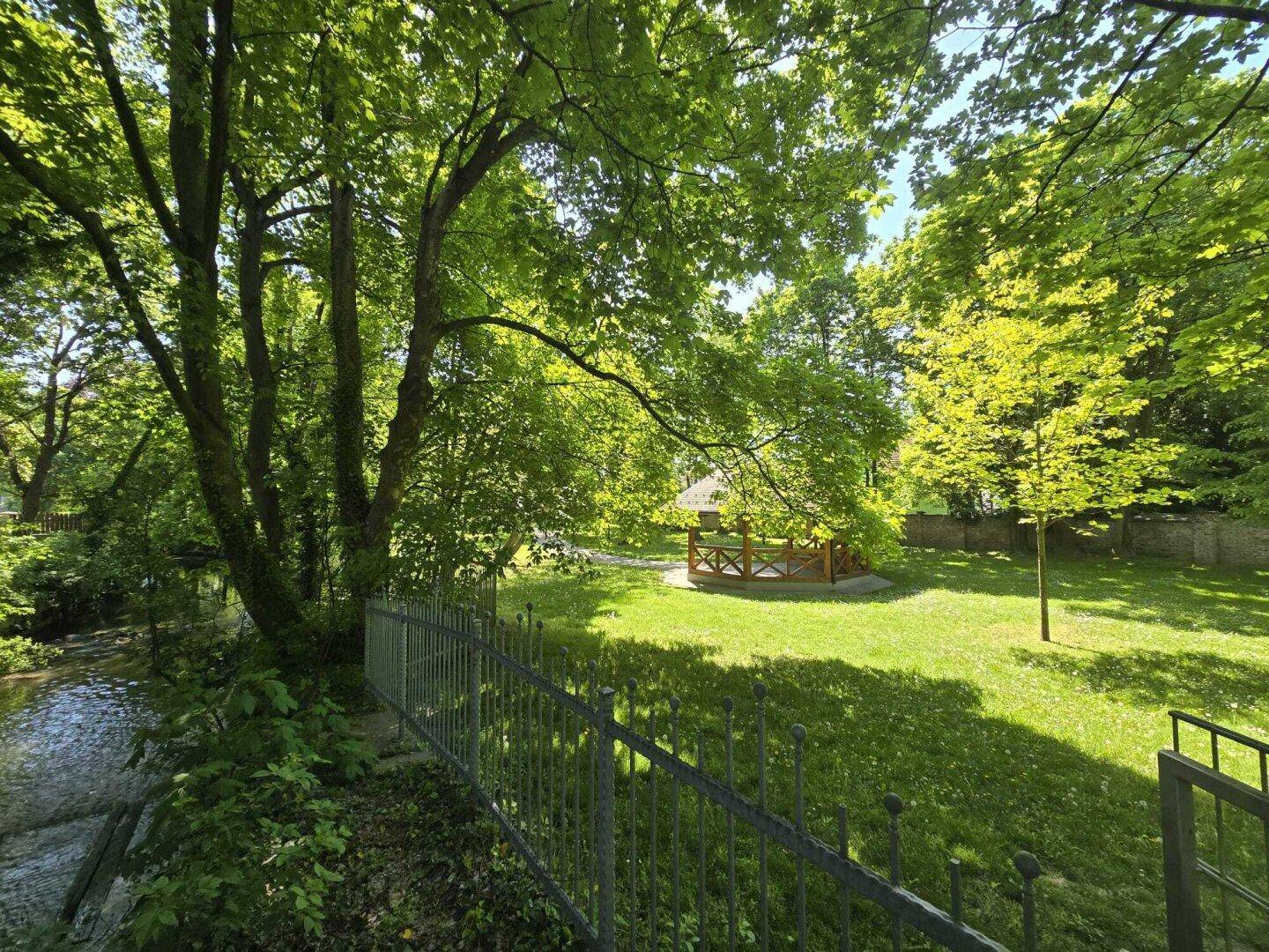 Grüne Gartenfläche am Flussufer mit einem kleinen Holzpavillon unter schattenspendenden Bäumen.
