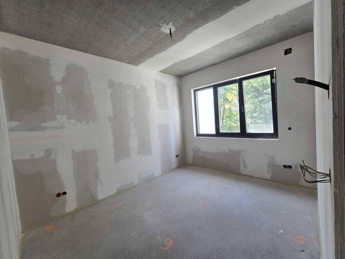 Raum im Rohbau mit verputzten Wänden und einem Fenster, das Tageslicht spendet.
