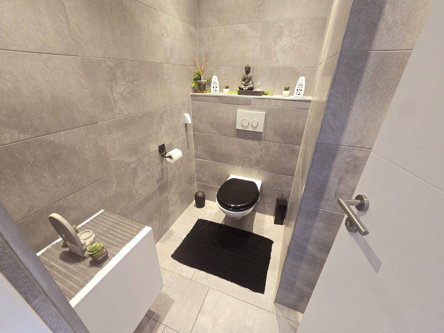 Modernes Gäste-WC mit grauen Fliesen, wandhängendem WC und kleinem Waschbeckenunterschrank.