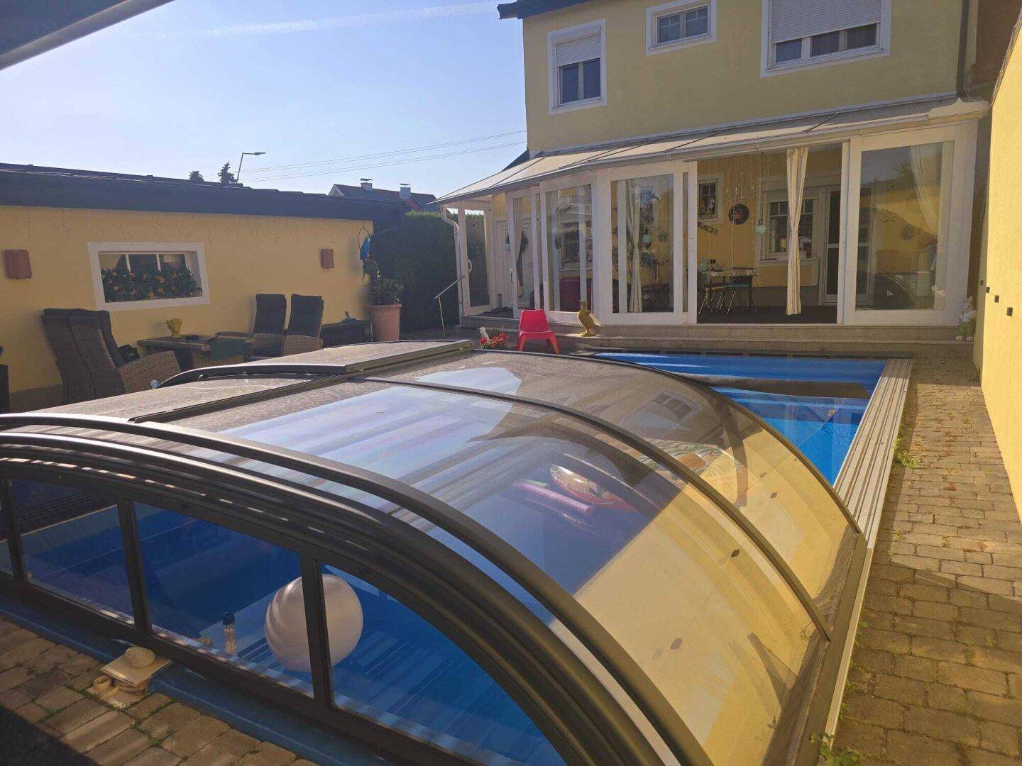 Hausfassade mit Poolbereich und Wintergarten unter blauem Himmel.