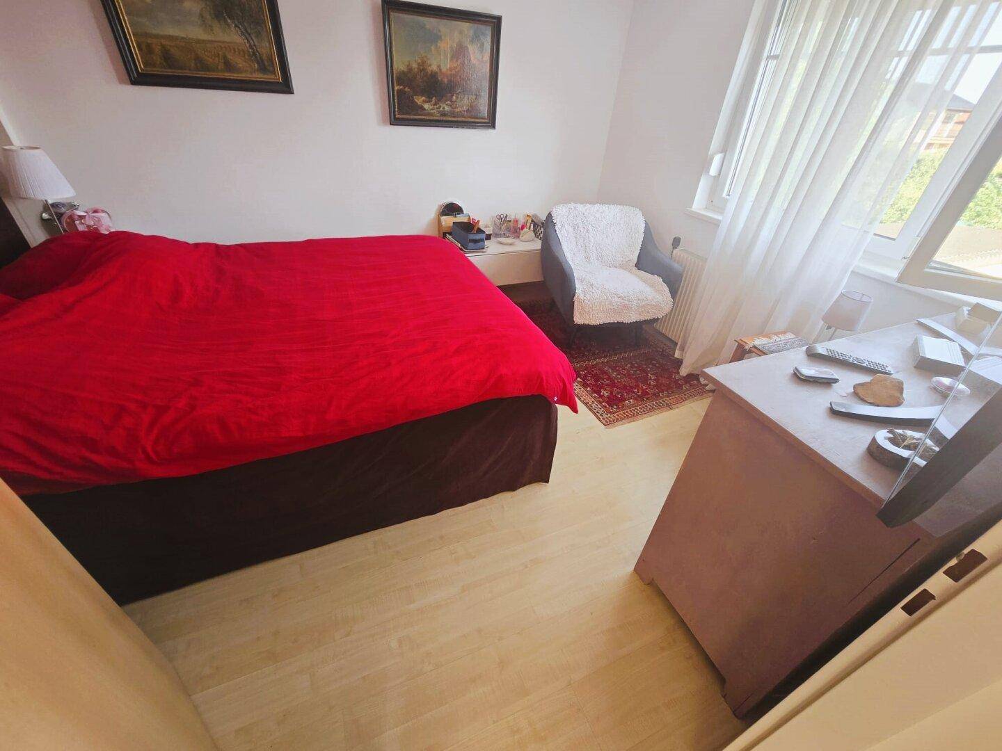 Helles Schlafzimmer mit roter Bettdecke, kleinem Sessel und Fenster, bietet viel Tageslicht.