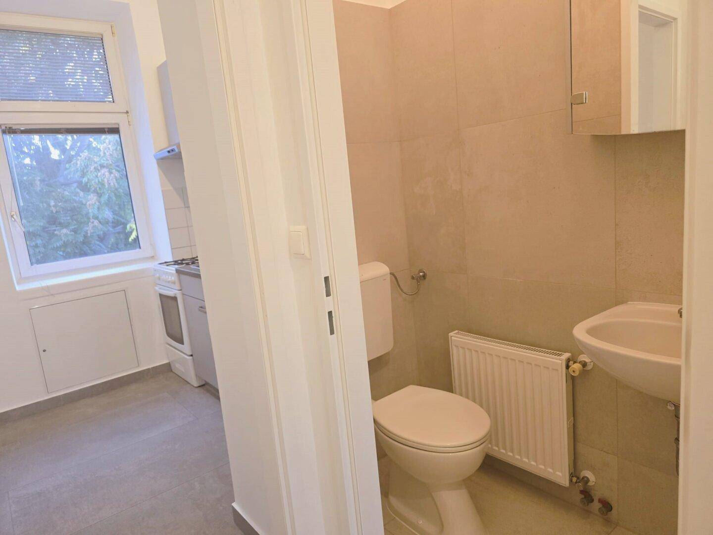 Kleines WC mit Toilette, Waschbecken und Heizkörper, mit Blick in die Küche.
