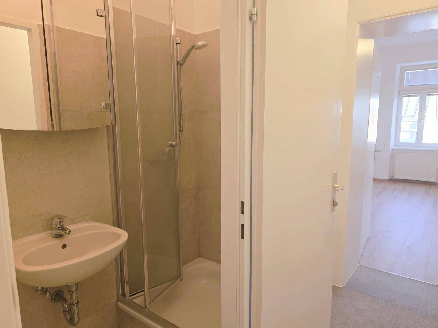 Kleines Badezimmer mit Duschkabine, Waschbecken und hellen Fliesen, mit Blick in einen Raum.