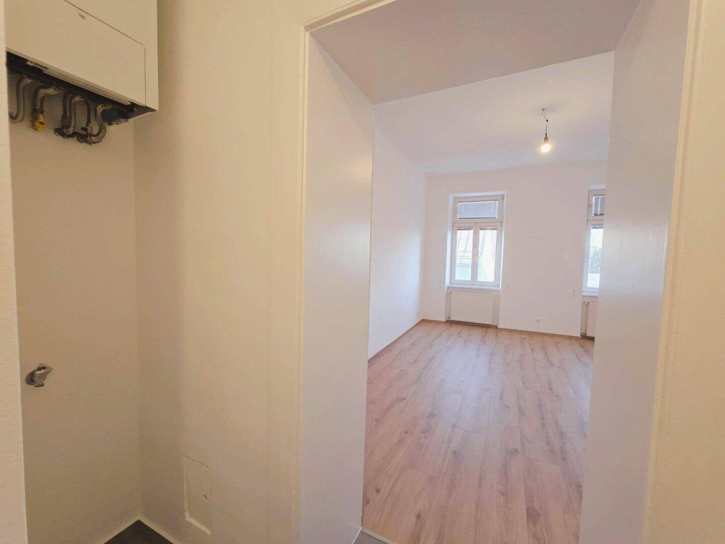Flur mit Heizungsanlage, der in ein helles Zimmer mit Laminatboden führt.