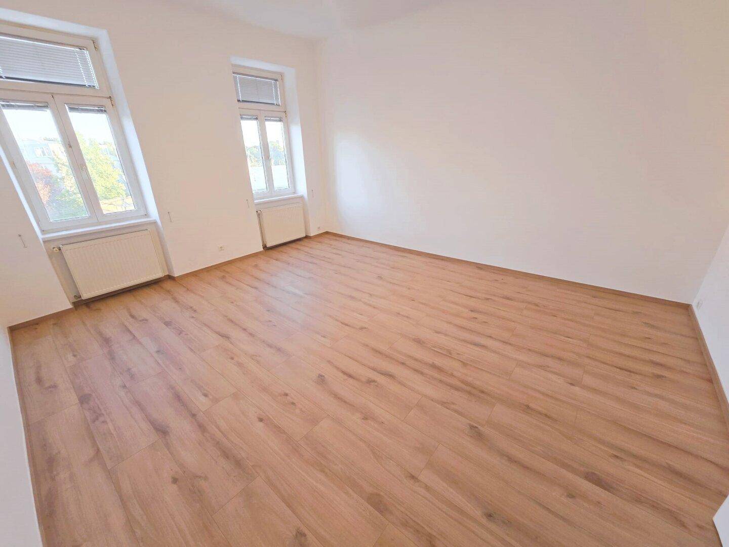 Helles, leeres Zimmer mit Laminatboden und zwei Fenstern, bereit zur Einrichtung.