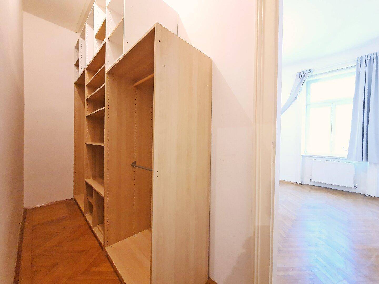 Heller Ankleidebereich mit offenen Regalsystemen und Blick in ein angrenzendes Zimmer mit Fenster.