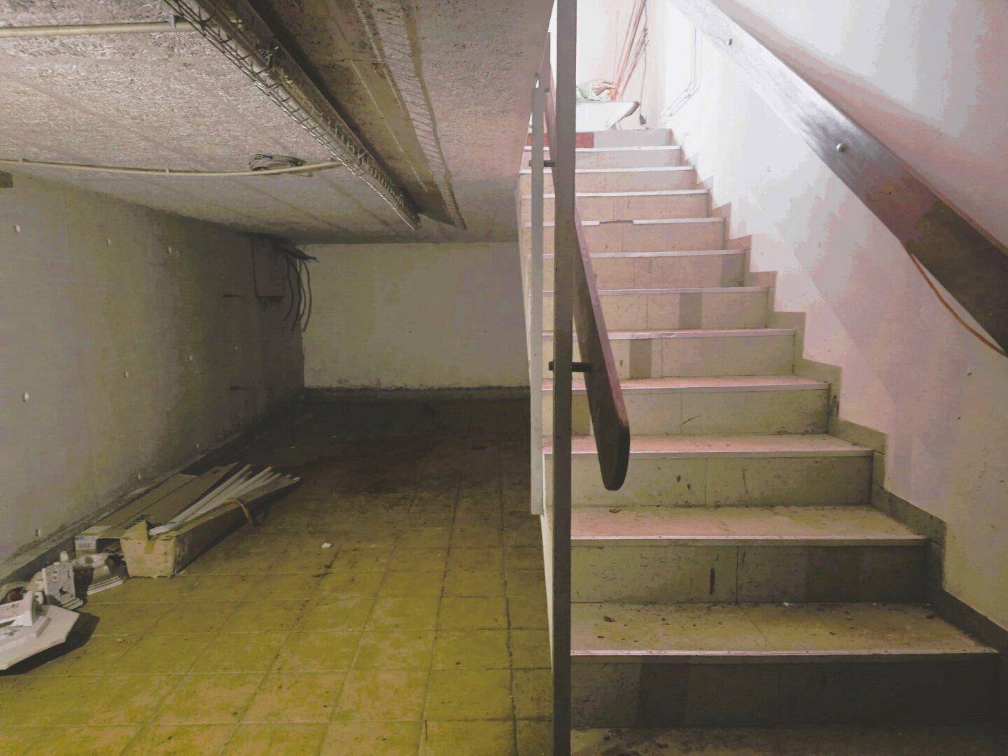 Treppenabgang in einen unfertigen Kellerraum mit einfacher Beleuchtung und Fliesenboden.