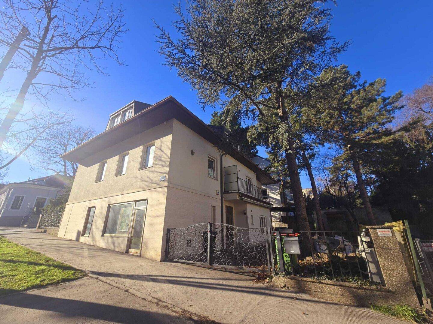 Das Haus präsentiert sich mit Balkon und schmiedeeisernem Tor, umgeben von hohen Bäumen.