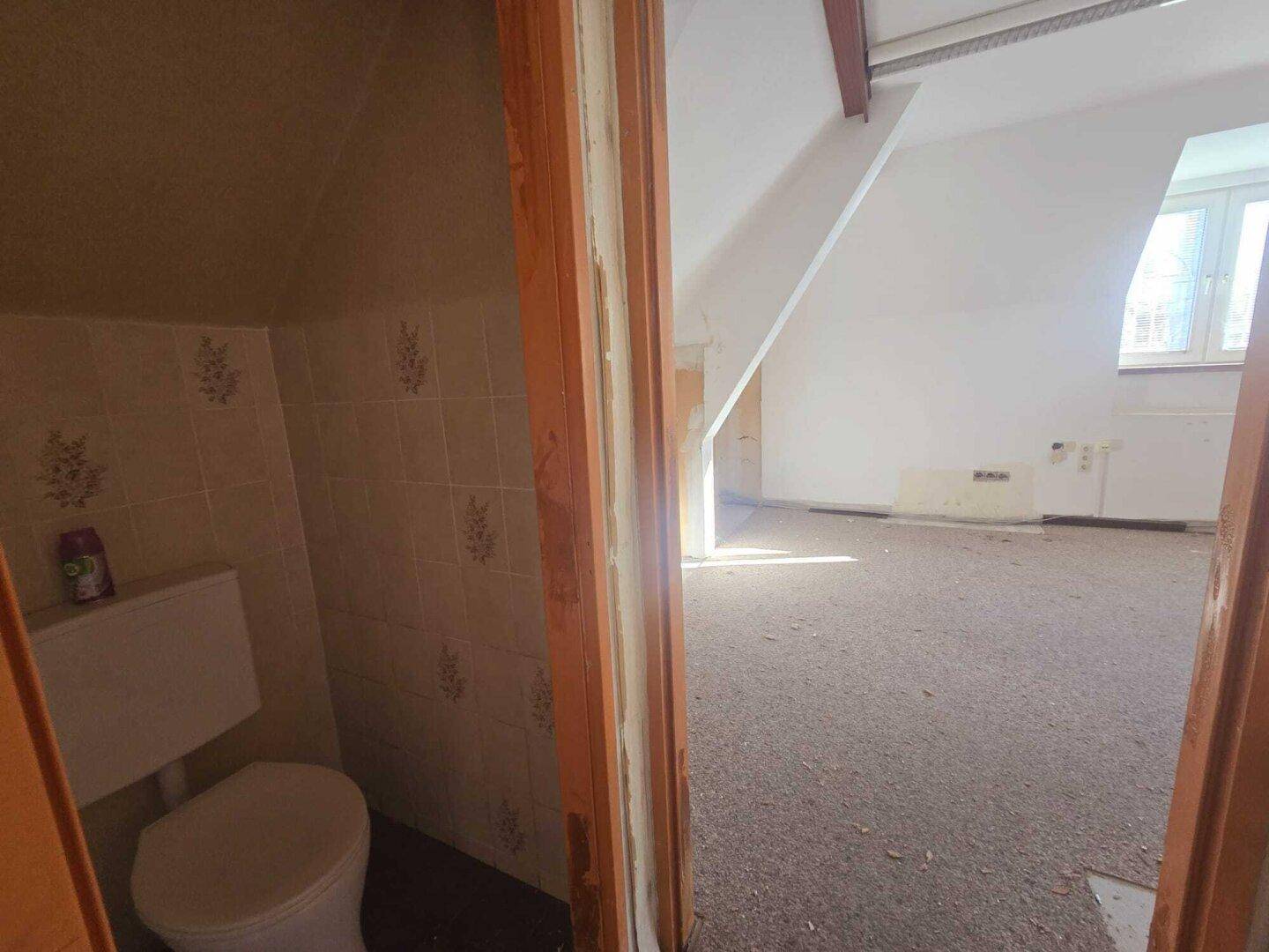 Kleines Gäste-WC mit Fliesen und Blick in einen angrenzenden, renovierungsbedürftigen Raum.