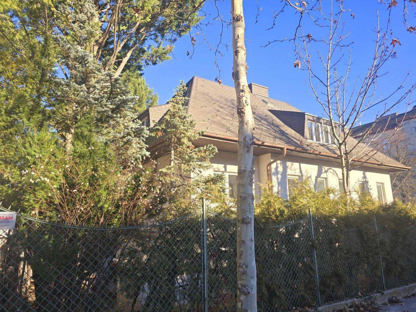 Ansicht des Hauses hinter einer Hecke und Bäumen mit Mansarddach unter blauem Himmel.