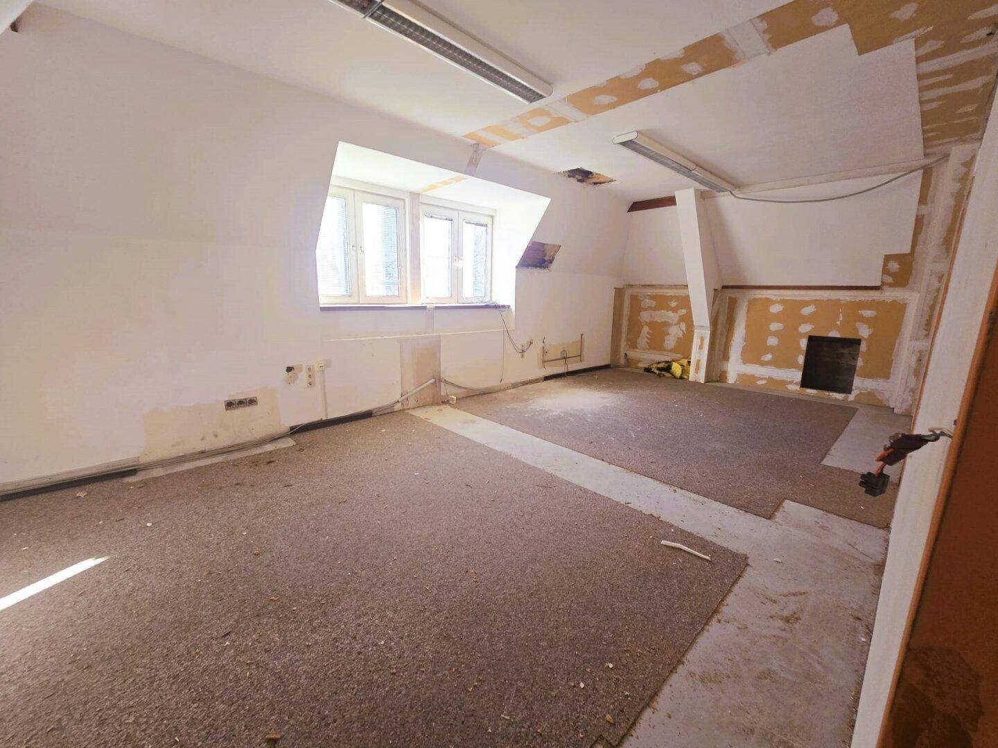 Renovierungsbedürftiger Raum im Dachgeschoss mit schrägen Wänden und Fenstern.