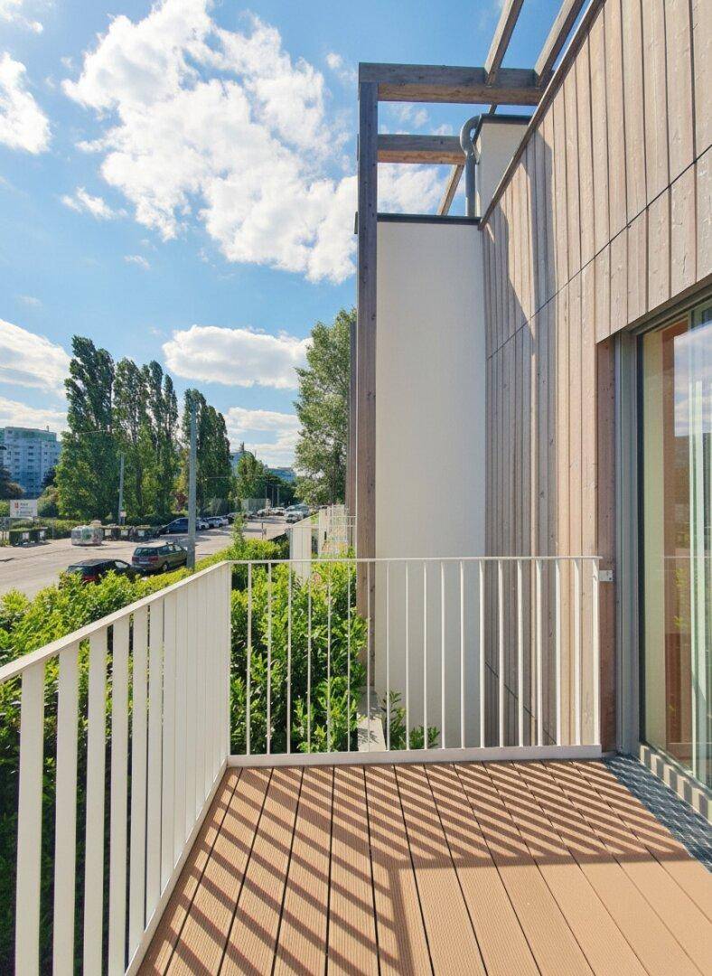 Balkon mit Holzbelag und weißem Geländer, umgeben von grünen Büschen.