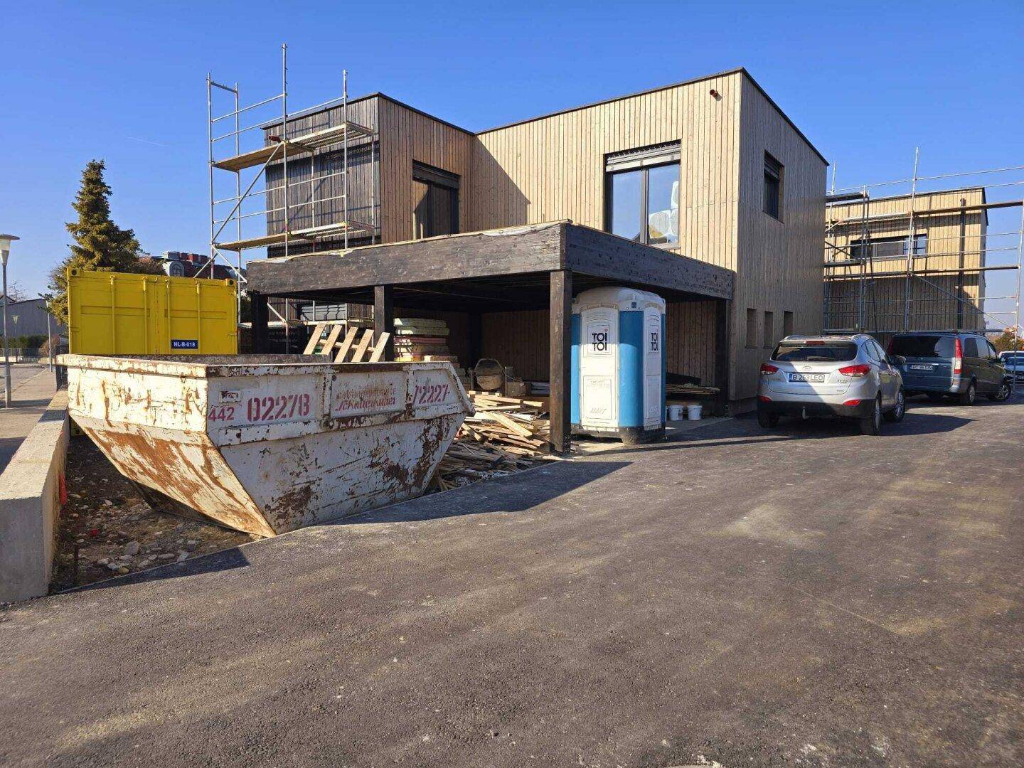 Neubau mit Holzfassade und Carport, umgeben von Baustellenmaterialien und Containern.