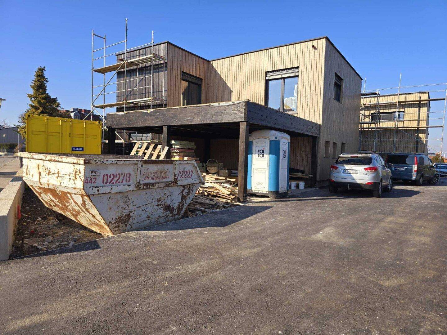 Neubau mit Holzfassade und Carport, umgeben von Baustellenmaterialien und Containern.