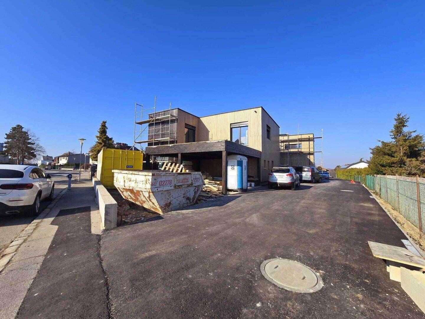 Gesamtansicht des Neubaus mit Holzfassade, Carport und Baustellenbereich an einem sonnigen Tag.