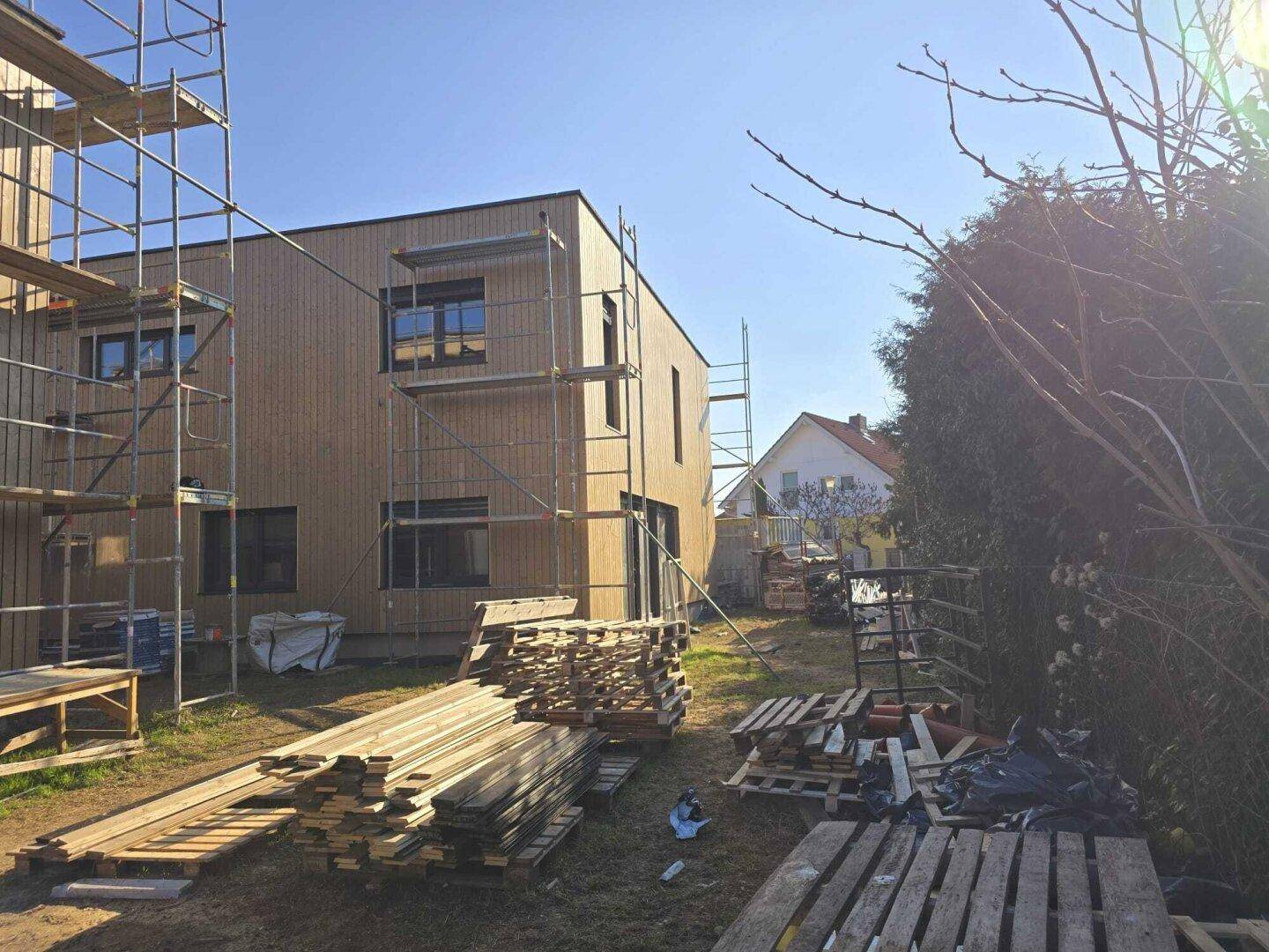 Hinterhofbereich eines Neubaus mit Holzfassade, Gerüst und Baumaterialien im Garten.