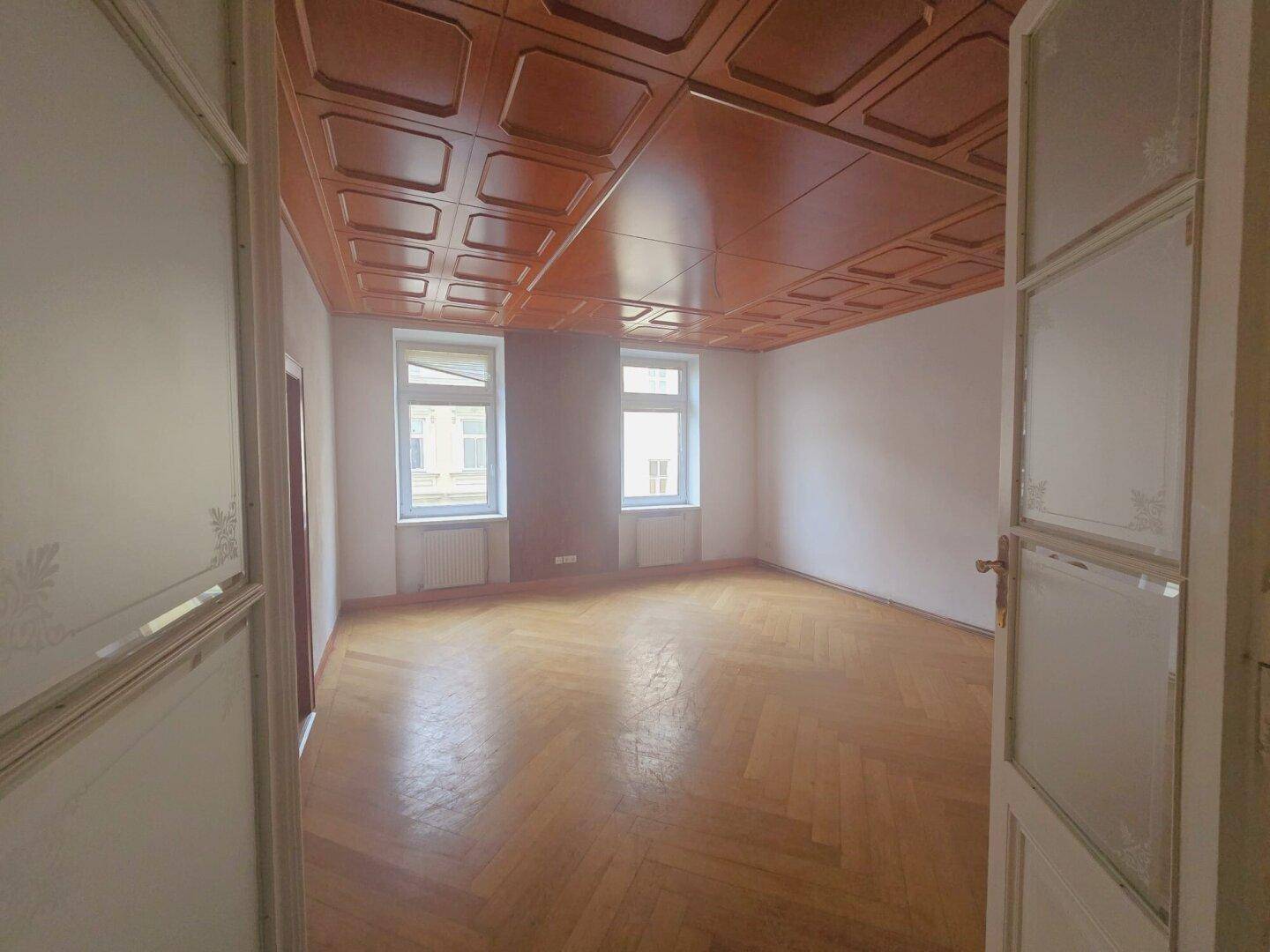 Helles Zimmer mit zwei Fenstern, Fischgrätparkett und dekorativer Holzdecke.