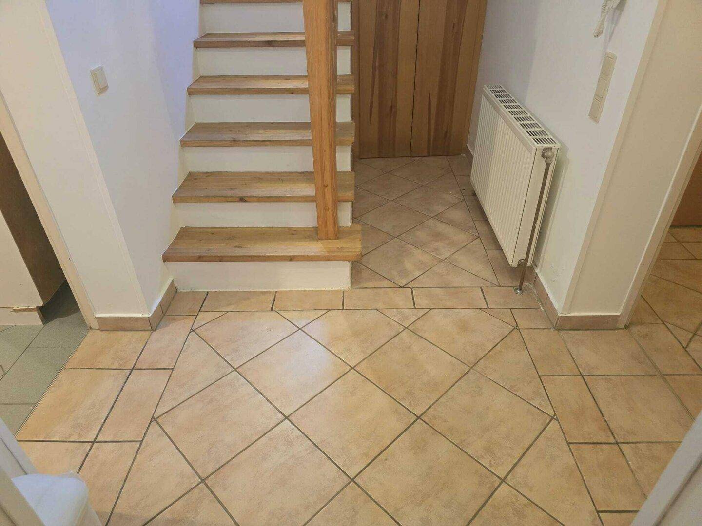 Eingangsbereich mit gefliestem Boden, Holztreppe und Heizkörper an der Wand.