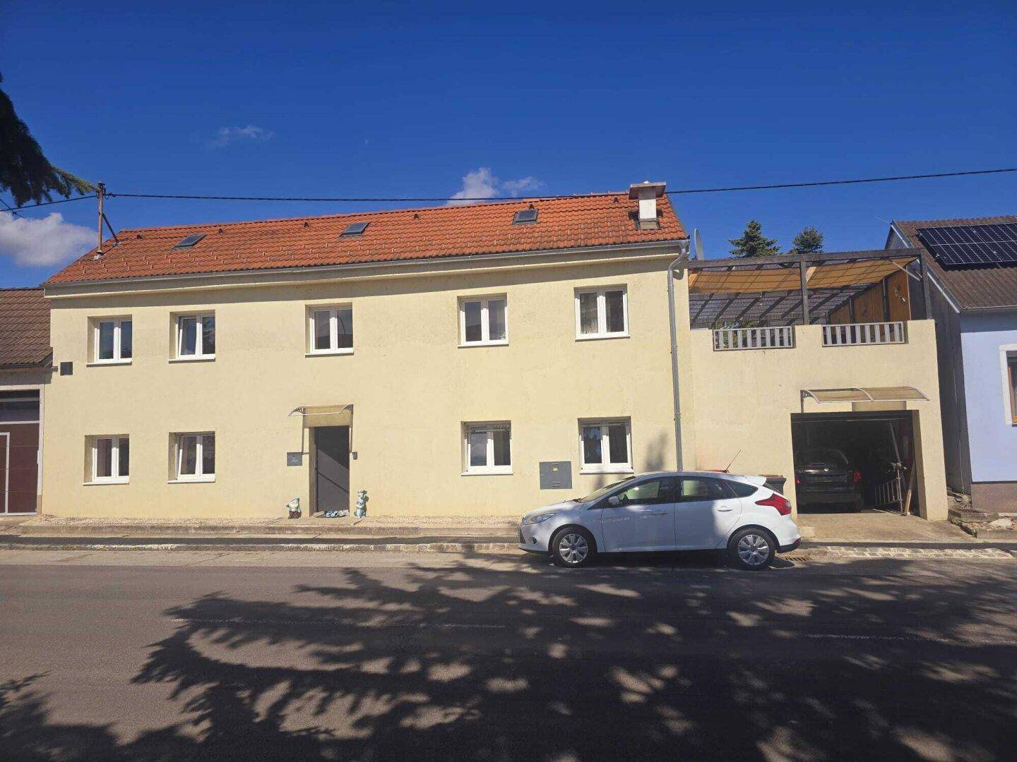Gelbe Fassade eines Wohnhauses mit rotem Ziegeldach, Garage und einer kleinen Terrasse im Obergeschoss.