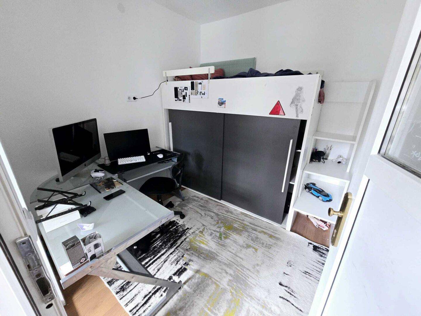 Jugendzimmer mit Hochbett, Schreibtisch und Computerarbeitsplatz, hell durch ein Fenster.