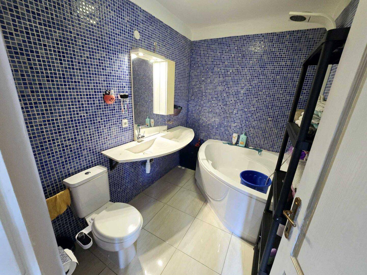 Badezimmer mit blauen Mosaikfliesen, Eckbadewanne, Toilette und einem einzelnen Waschbecken.