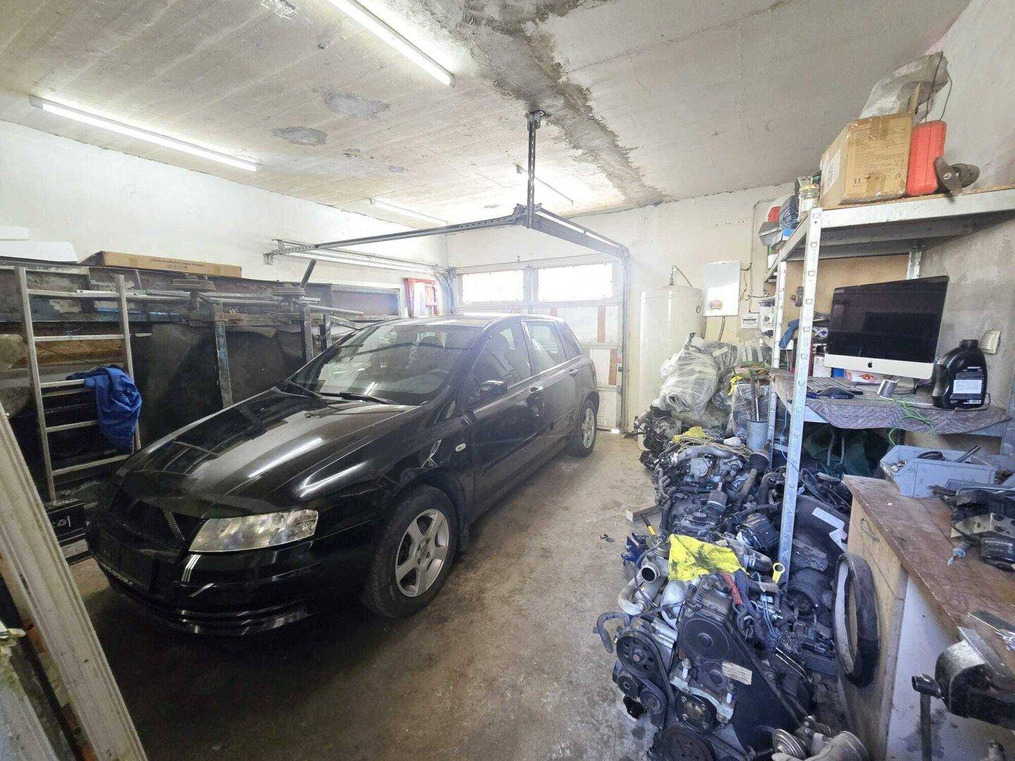 Geräumige Garage mit einem Auto, Werkzeugen und Ersatzteilen, beleuchtet durch Neonröhren.