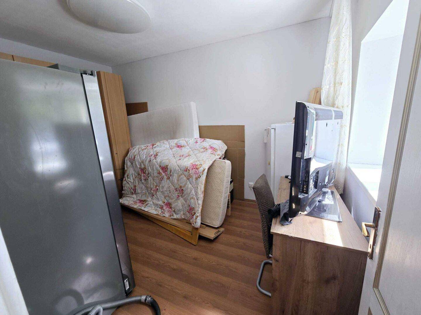Unaufgeräumter Raum mit Laminatboden, Fenster und verschiedenen Möbelstücken und Geräten.