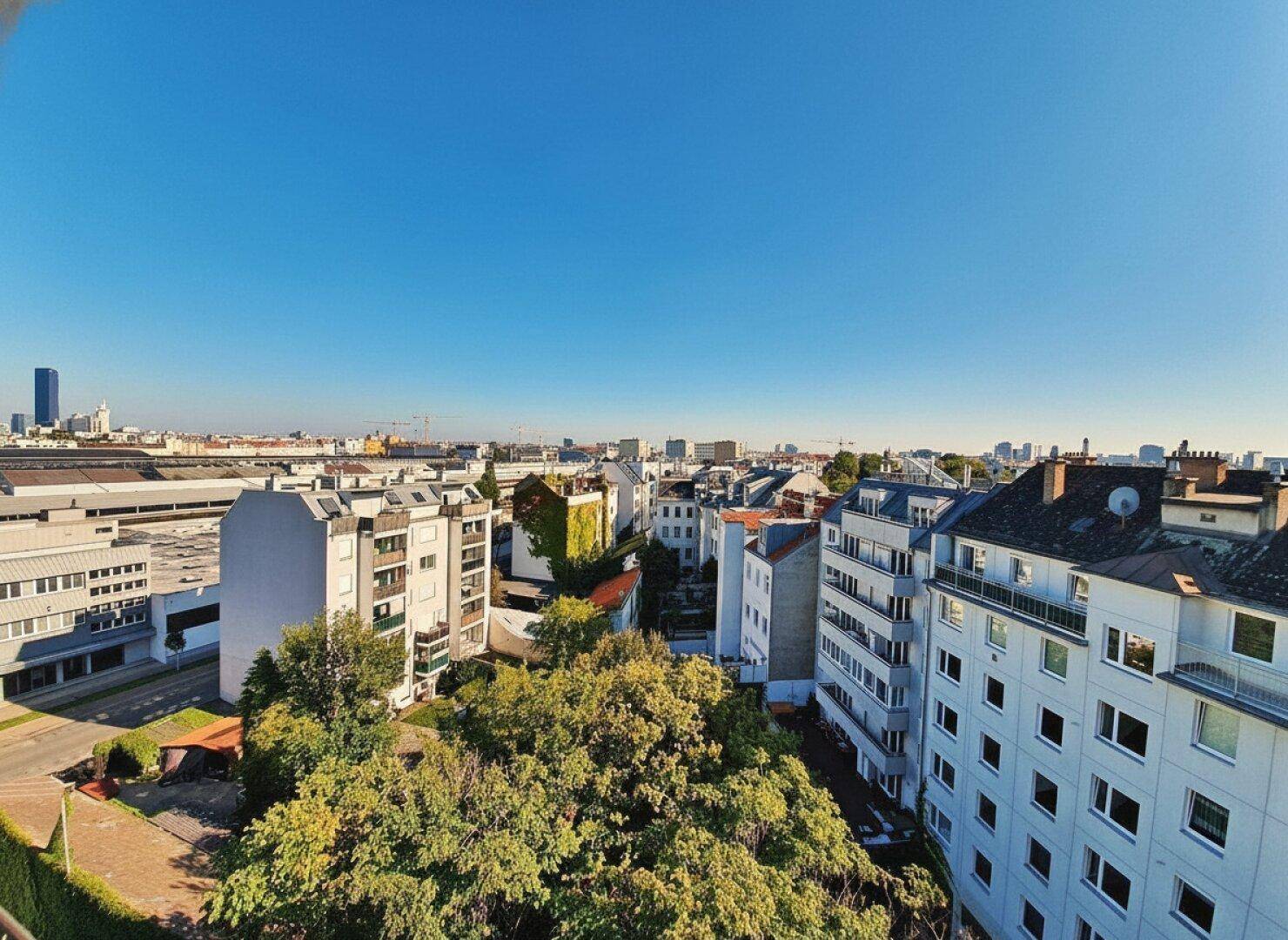 Breiter Ausblick auf die städtische Umgebung mit Gebäuden, Bäumen und einem klaren blauen Himmel.
