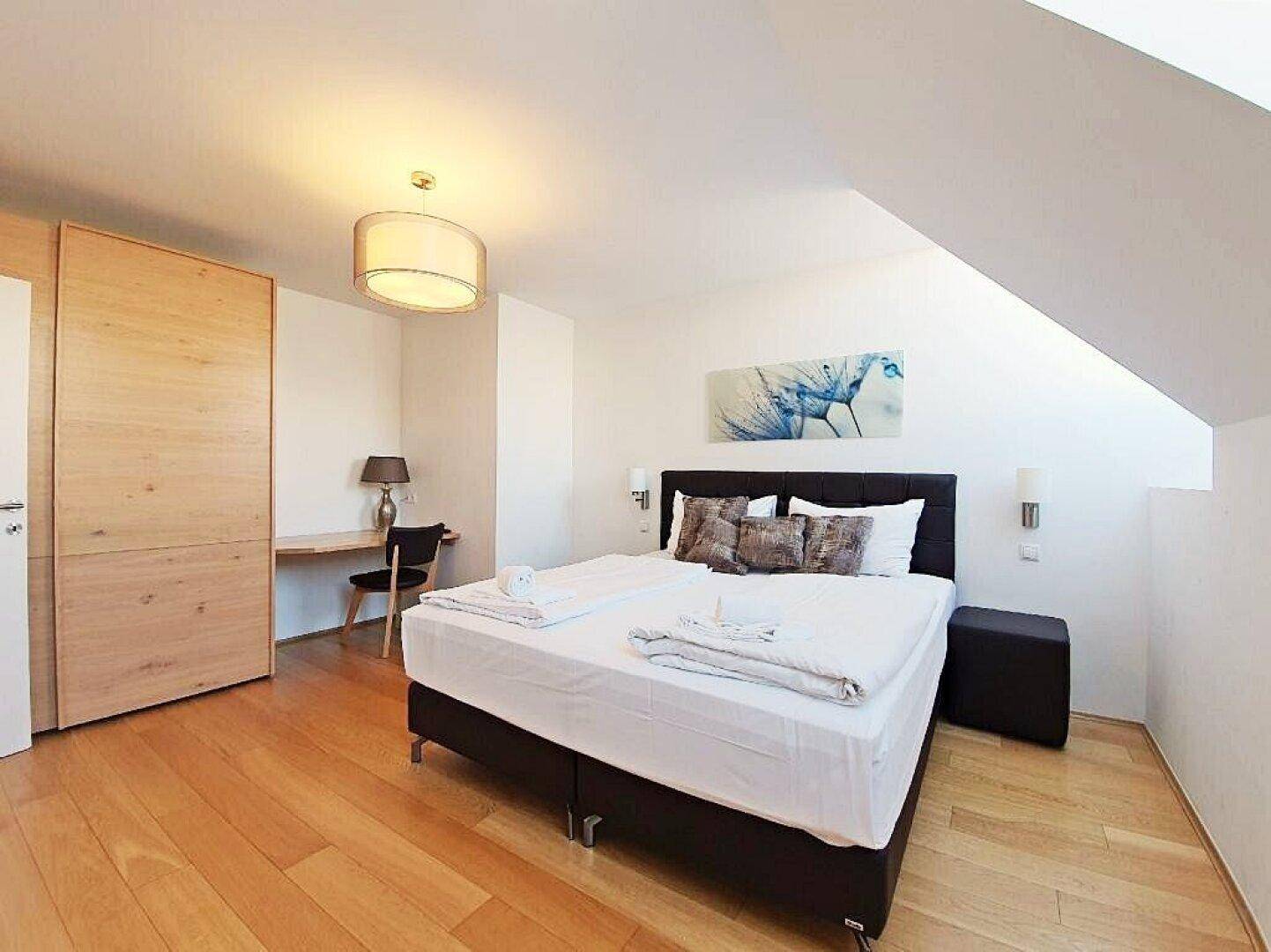 Schlafzimmer mit Doppelbett, Holzboden, Einbauschrank und einem kleinen Arbeitsbereich unter der Dachschräge.