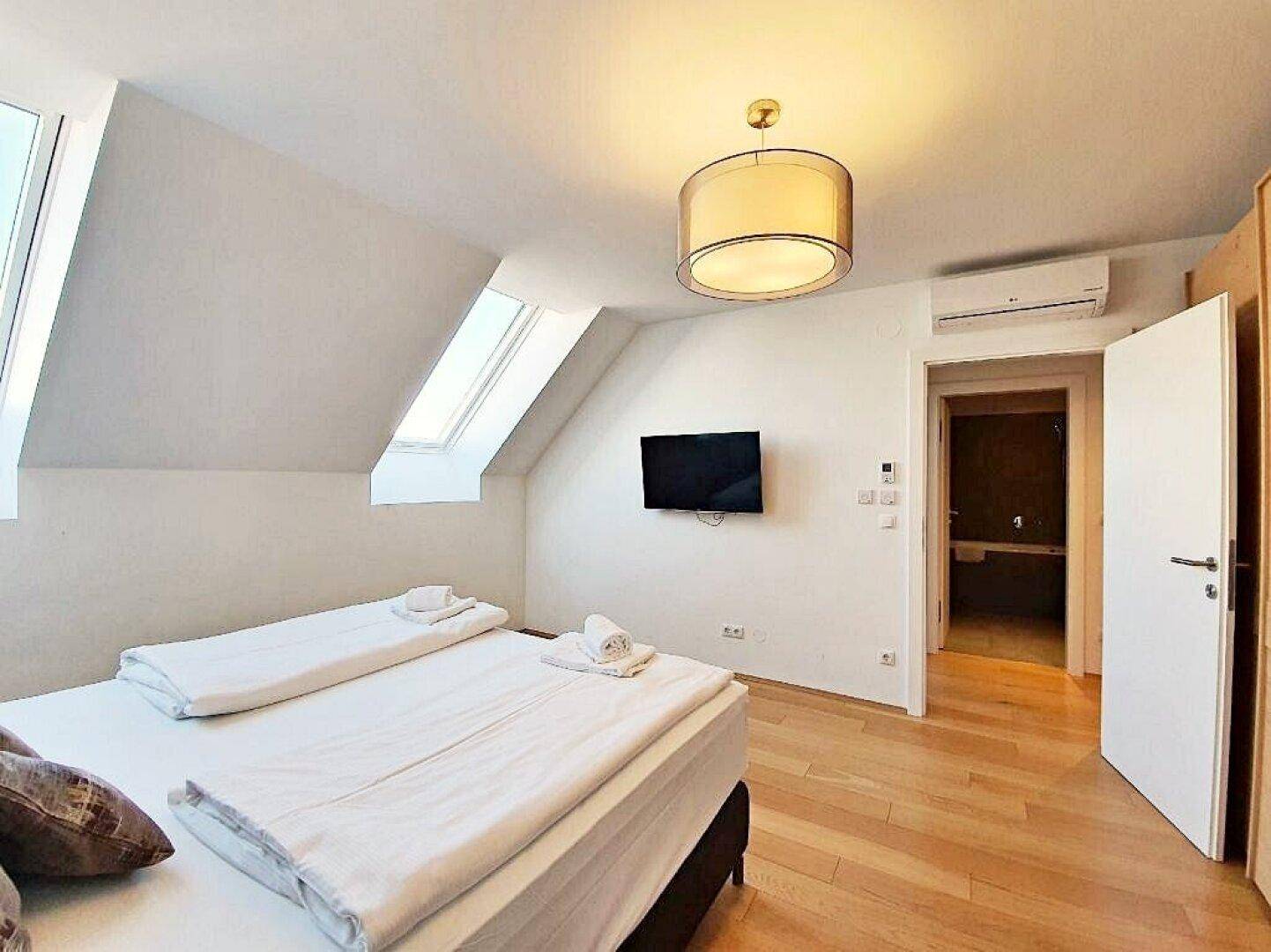 Schlafzimmer mit Doppelbett, Wandfernseher und Zugang zu einem angrenzenden Badezimmer, inklusive Klimaanlage.